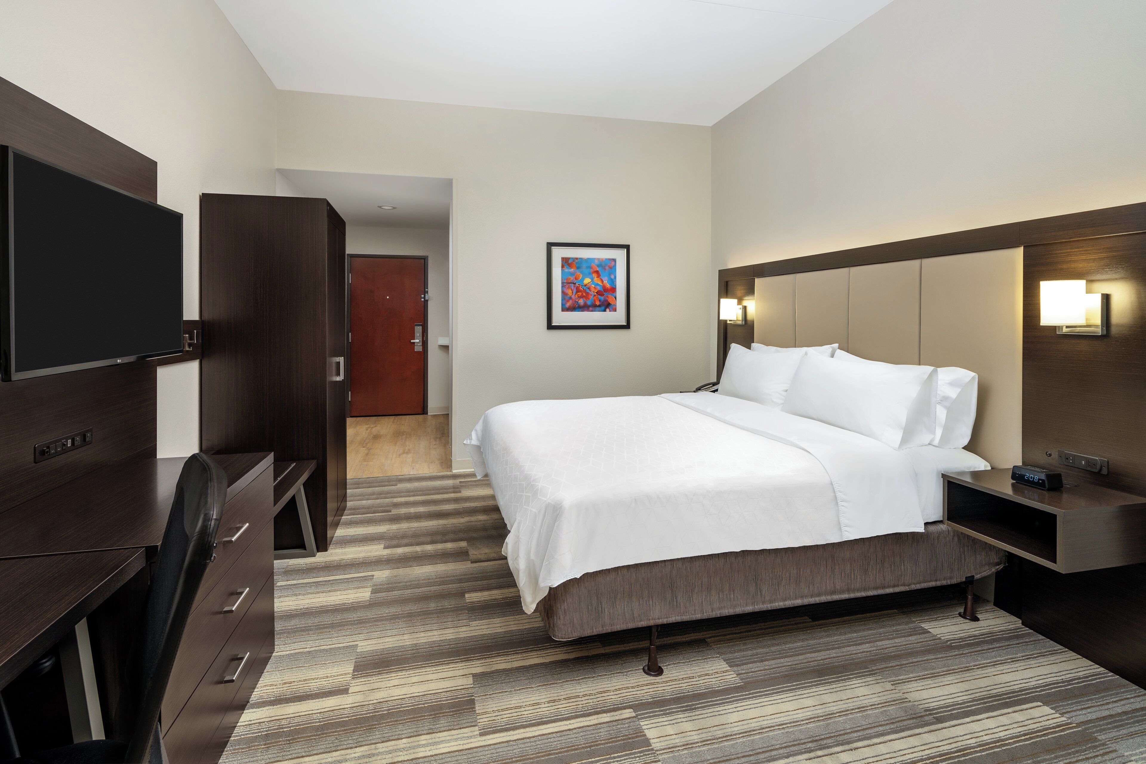 A hotel room with a bed, a desk, a TV, and a painting on the wall.