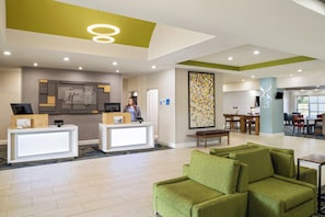 A hotel lobby with a reception desk, a seating area with green sofas, and a dining area with tables and chairs.