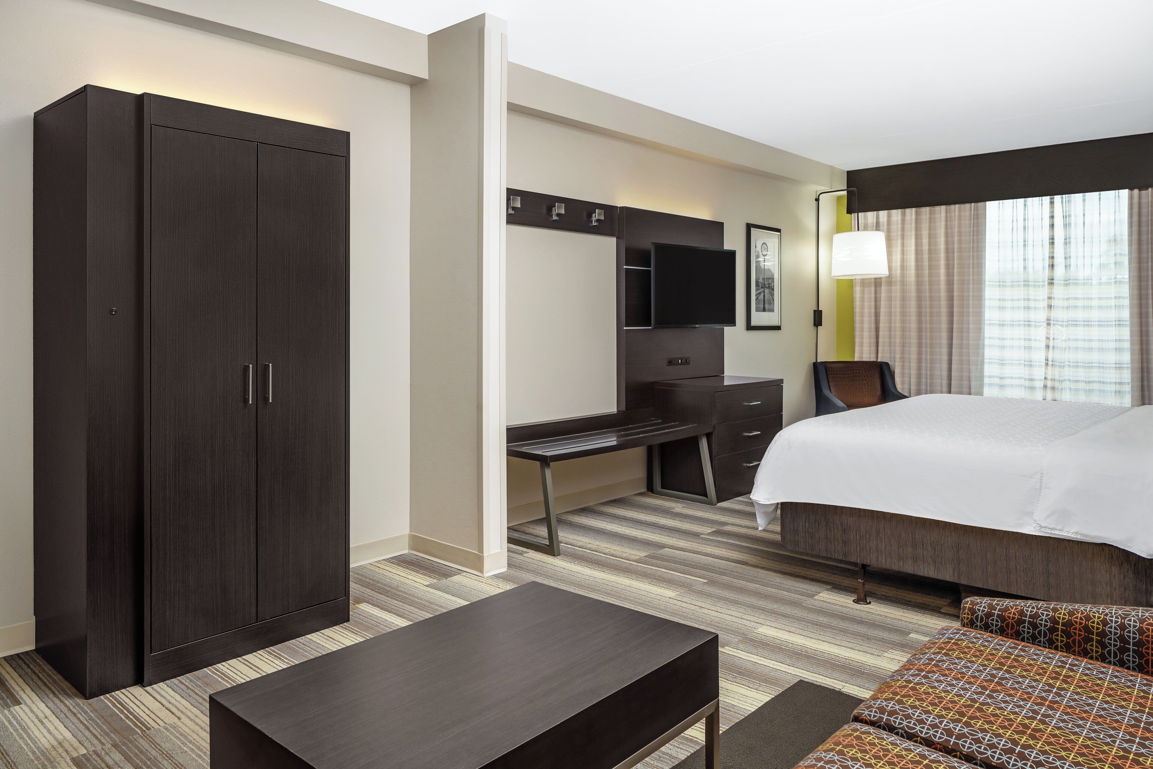 Quarto de hotel com cama, escrivaninha, guarda-roupa, televisão e janela com cortinas.