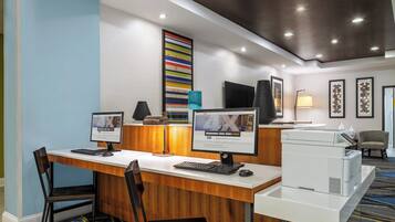Zona de recepción de un hotel con dos puestos de trabajo de ordenador, un mostrador de recepción, un televisor de pantalla plana, una impresora y un área de descanso con una silla.