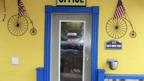 Un edificio amarillo con una puerta azul, un letrero que dice 'OFFICE' encima y banderas estadounidenses colgadas en la pared.