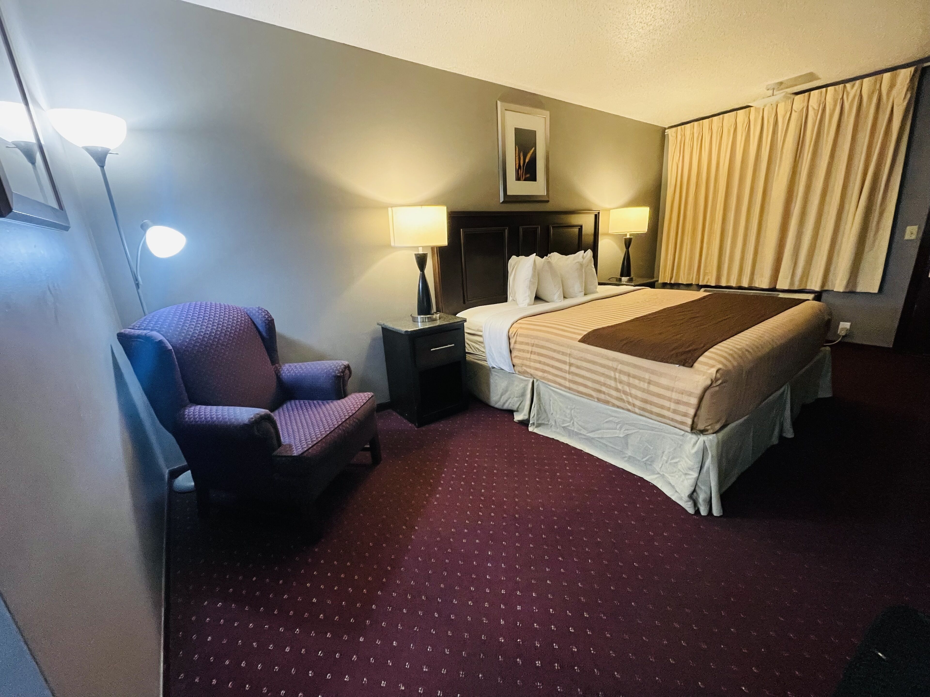 Une chambre d’hôtel avec un lit, une chaise, deux tables de chevet avec des lampes, un tableau au mur et une fenêtre avec des rideaux.