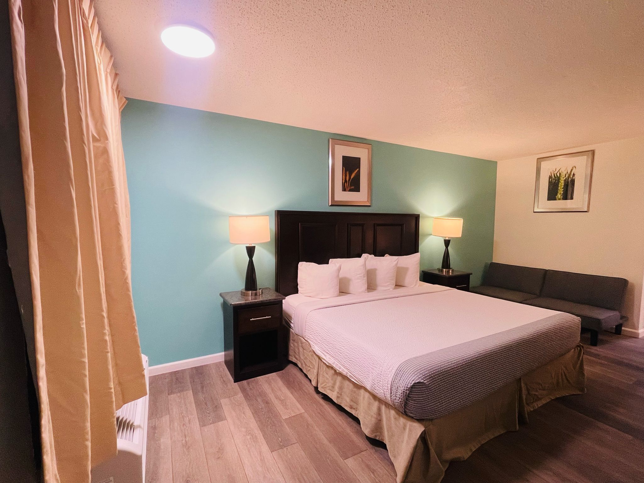 Una habitación de hotel con una cama, dos mesitas de noche con lámparas, un cabecero oscuro, un sofá gris y un cuadro enmarcado en la pared.