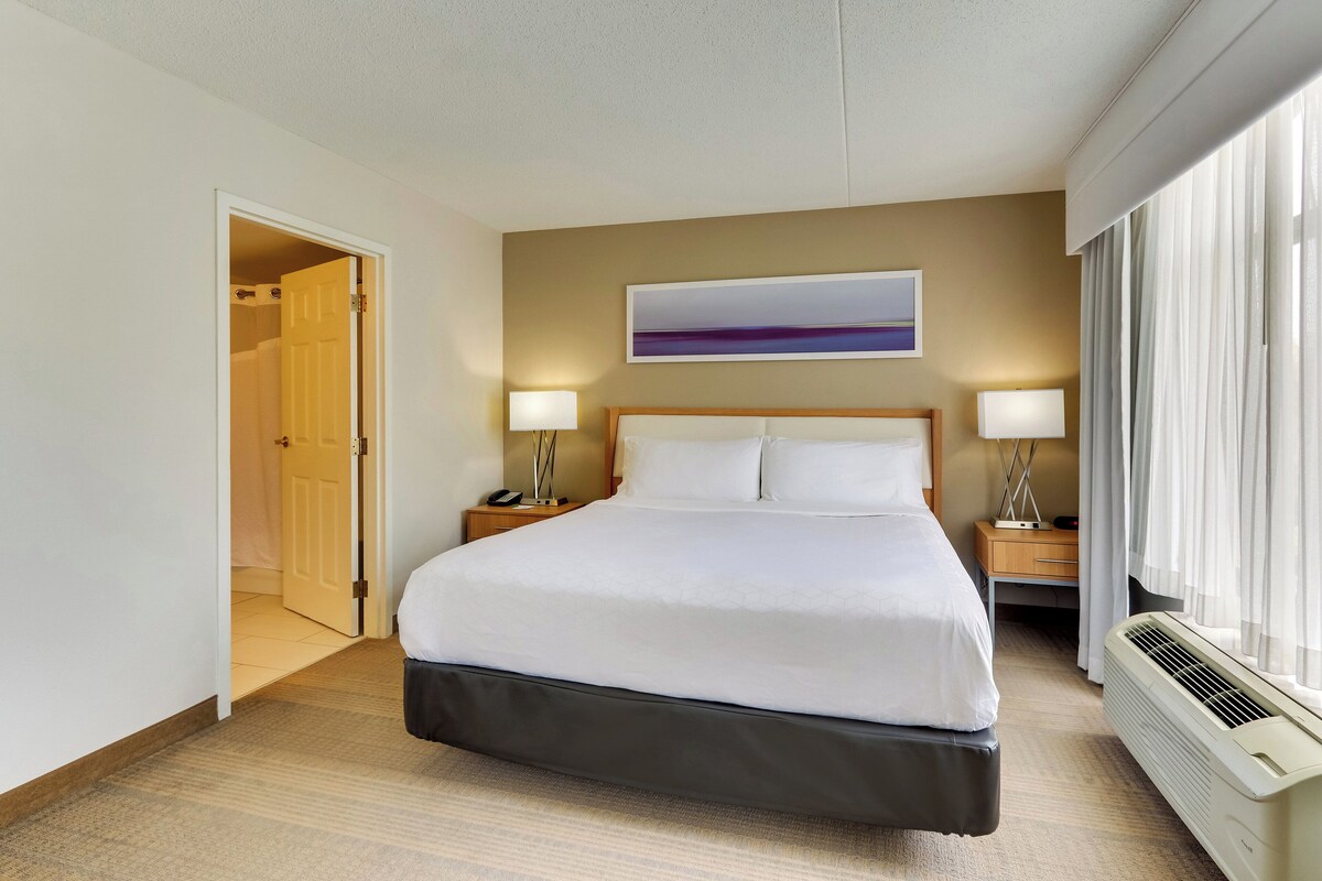 Habitación de hotel con una cama, lámparas de noche, un cuadro, una ventana con cortinas y una puerta que da a otra habitación.