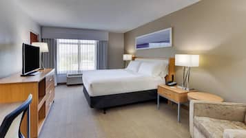 A hotel room with a bed, a desk with a TV, a chair, a bedside table with a lamp, and a window with curtains.