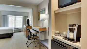 Habitación de hotel con cama, escritorio, silla, lámpara, microondas, cafetera y ventana con cortinas.