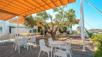 Un patio al aire libre con sillas y mesas blancas, cubierto por toldos naranjas, rodeado de árboles.