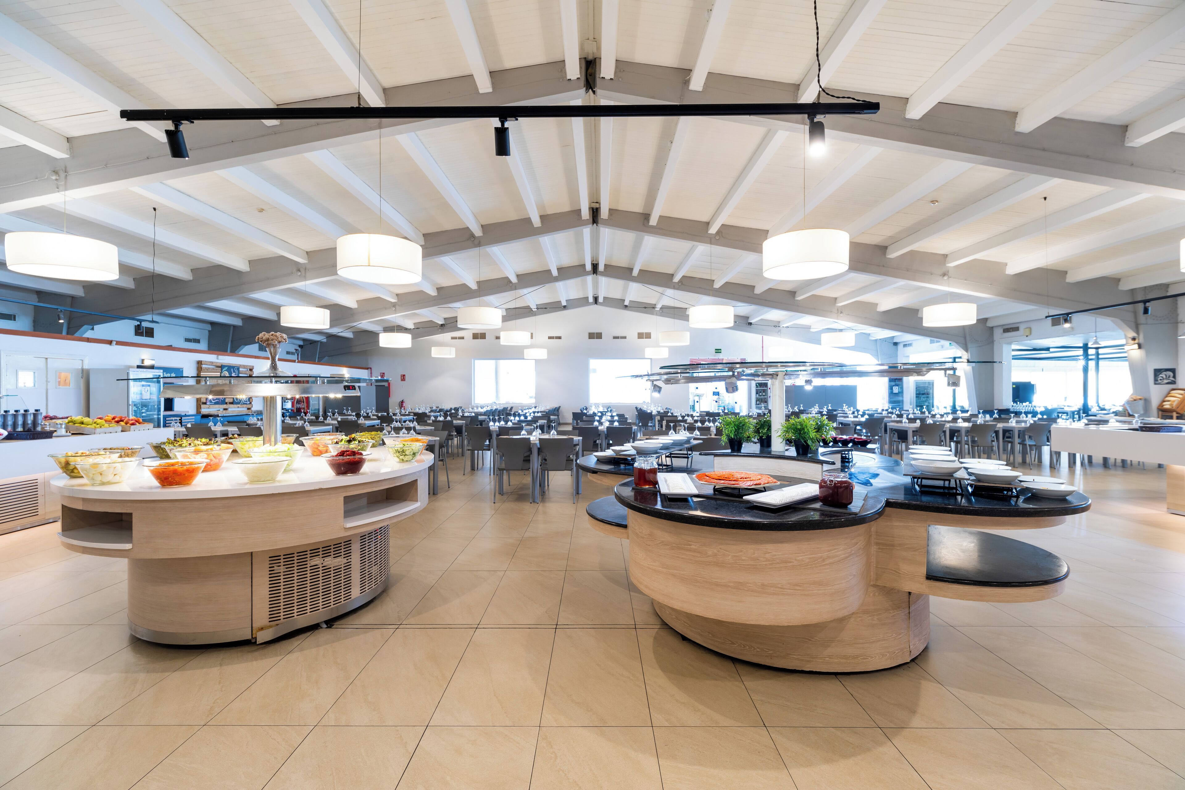 Una cafetería espaciosa con varios mostradores de servicio y mesas, techos altos con vigas a la vista y amplias ventanas que dejan entrar luz natural.