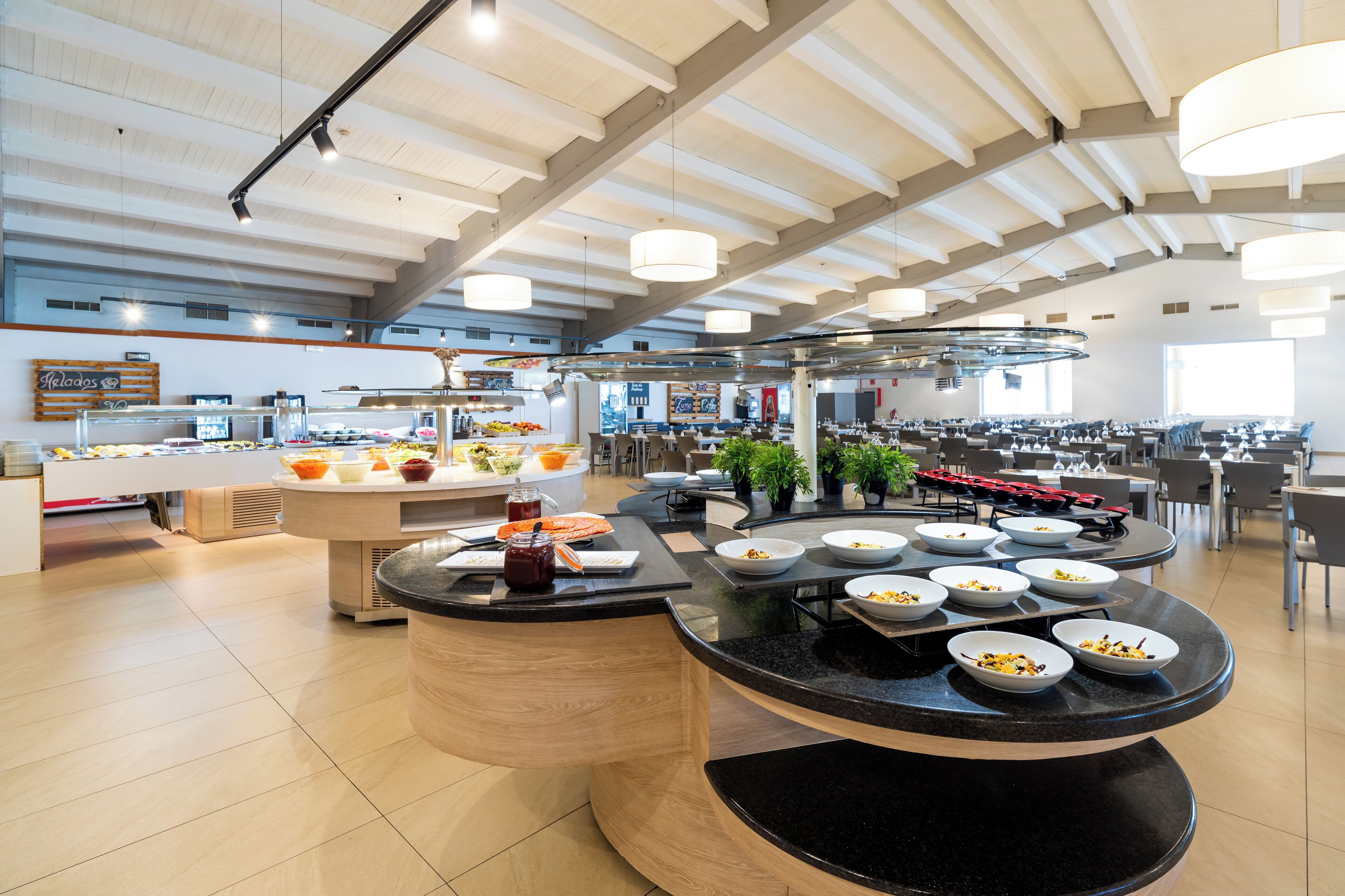 Une cafétéria moderne dotée de nombreuses tables, d’un espace de service central et d’un plafond haut avec des poutres apparentes.
