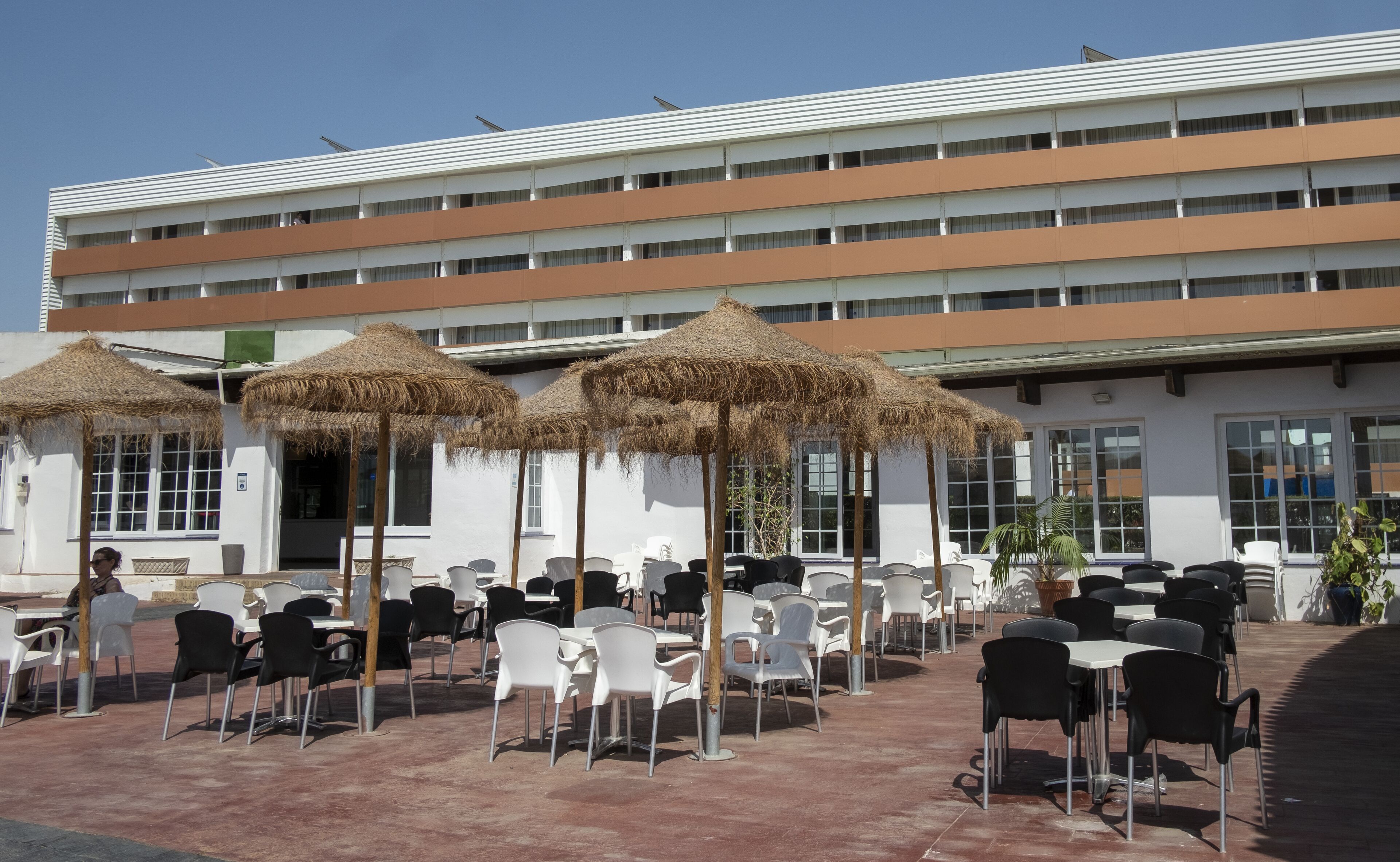 Un área de estar al aire libre con mesas y sillas, algunas con sombrillas de paja, y un edificio de varios pisos al fondo.
