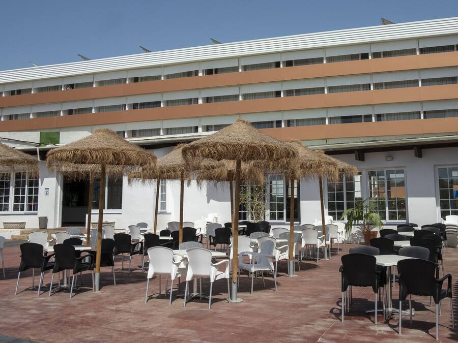 An outdoor seating area with tables and chairs, some with thatched umbrellas, and a building with multiple floors in the background.