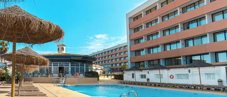 A hotel pool area with sun loungers and umbrellas, a swimming pool, and a building with multiple balconies.