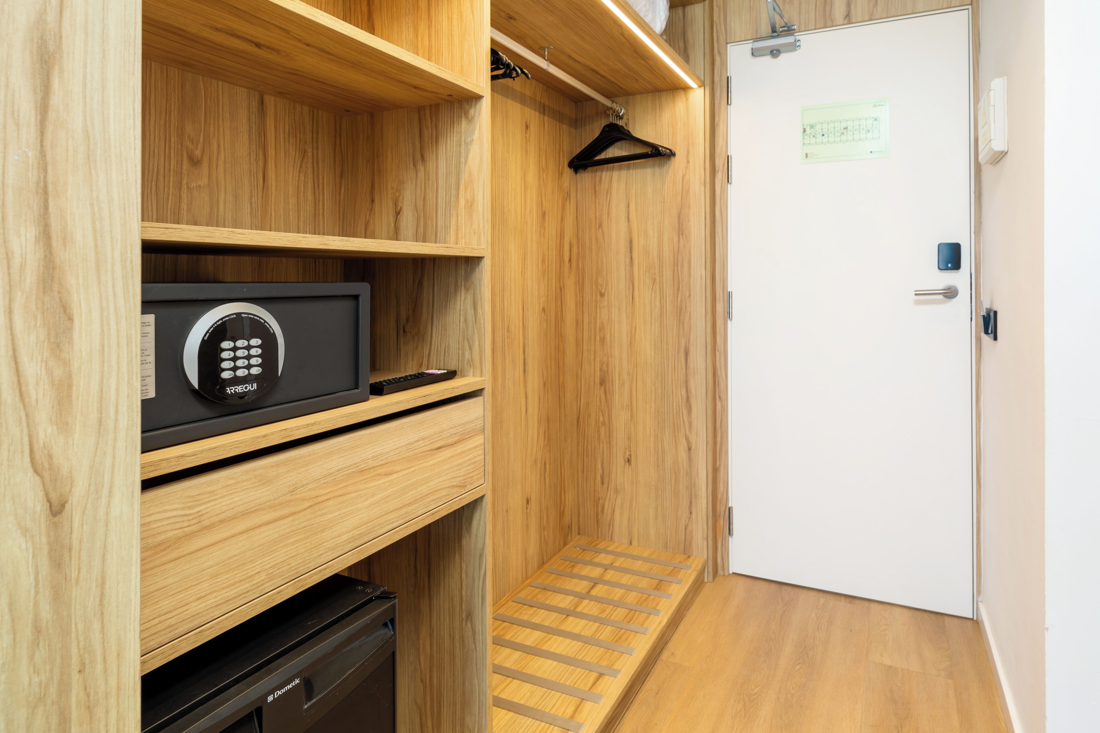 Una habitación pequeña con un armario de madera, una caja fuerte negra y una puerta blanca con un aviso.