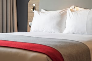 Un lit bien fait, avec du linge de lit blanc et une couverture rouge, une tête de lit en bois et un luminaire fixé au mur.