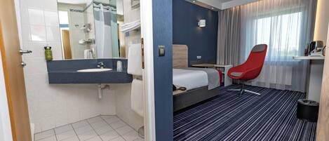 Una habitación de hotel con baño, una cama, una silla roja y una ventana con cortinas.