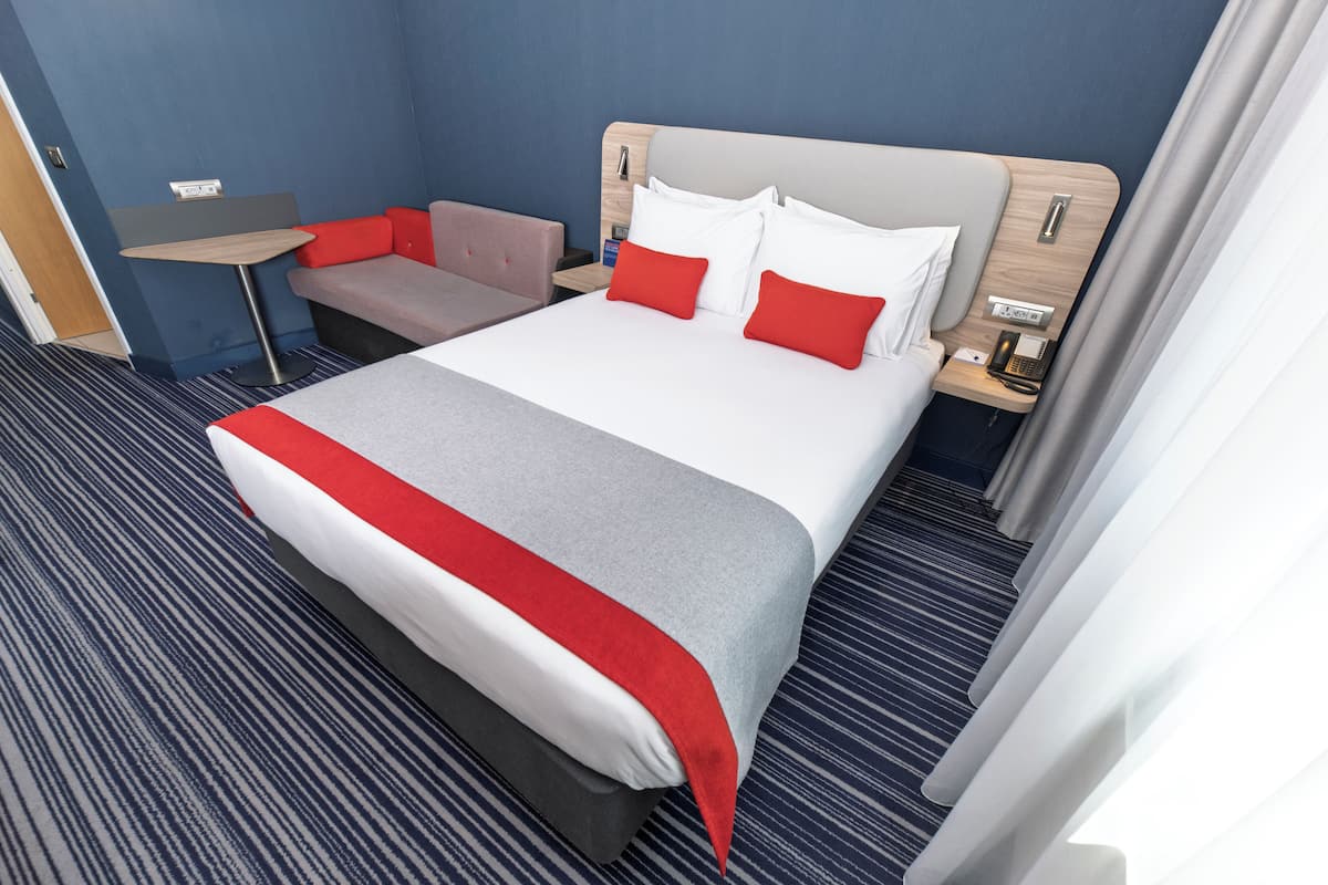 A hotel room with a large bed, a small desk, a sofa, and a nightstand with a phone.