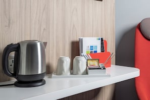 Un comptoir de cuisine avec une bouilloire, deux tasses, une machine à café, un porte-magazines et une chaise rouge.
