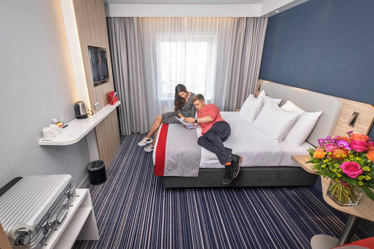 A hotel room with a bed, a bedside table, a small desk, a chair, a trash bin, and a window with curtains.