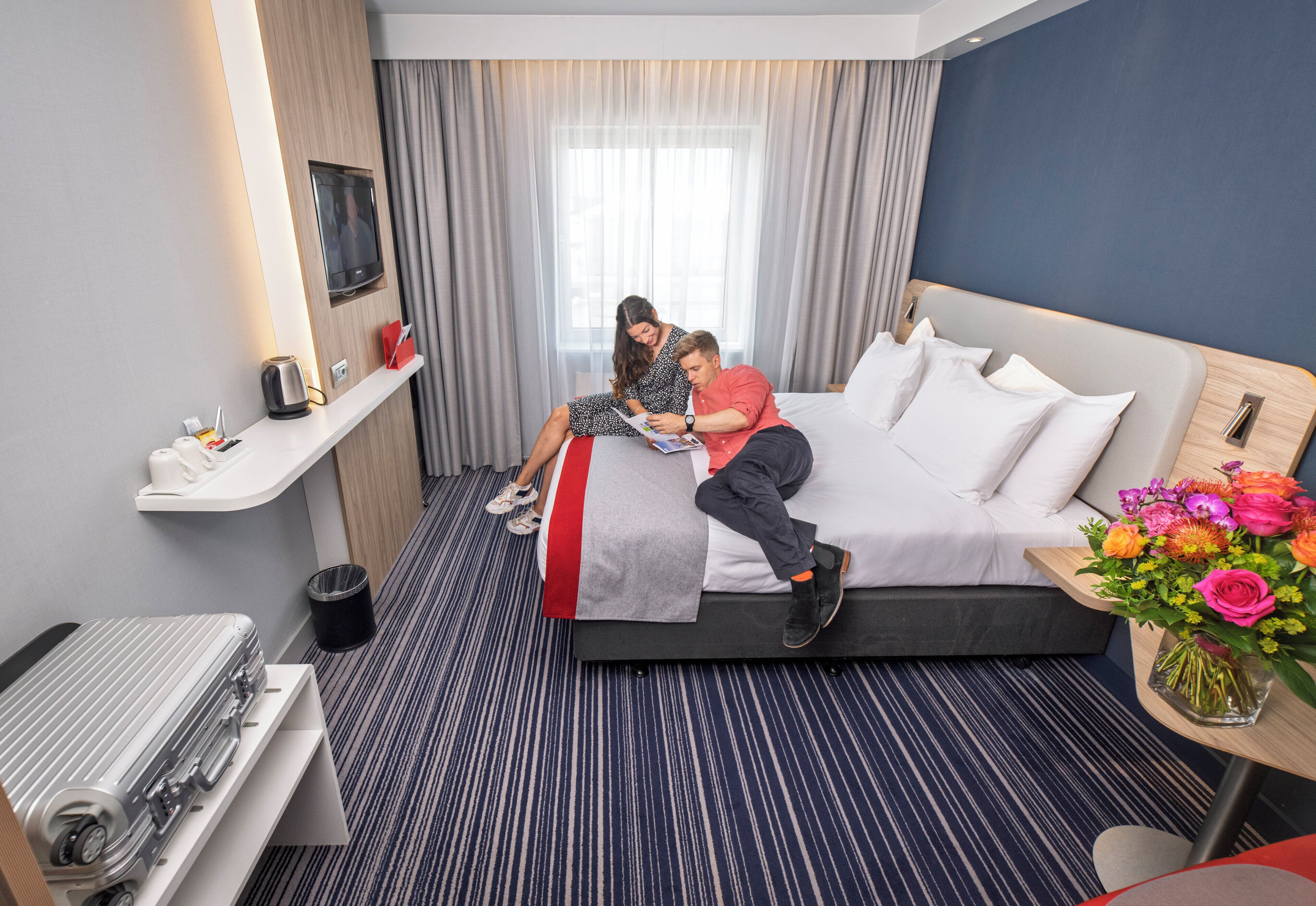 A hotel room with a bed, a bedside table, a small desk, a chair, a trash bin, and a window with curtains.