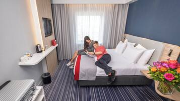 A hotel room with a bed, a bedside table, a small desk, a chair, a trash bin, and a window with curtains.