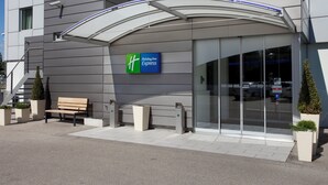 A modern building with a Holiday Inn Express sign, a covered entrance, a bench, and potted plants. - Holiday Inn Express Geneva Airport by IHG (Meyrin)