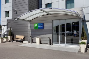 Moderni rakennus, jossa on Holiday Inn Express -kyltti, katettu sisäänkäynti, penkki ja ruukkukasveja.