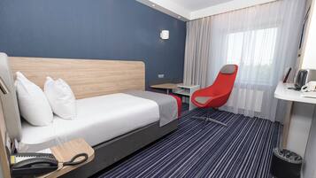 A hotel room with a bed, a desk, a red chair, a small table, a telephone, and a trash bin.