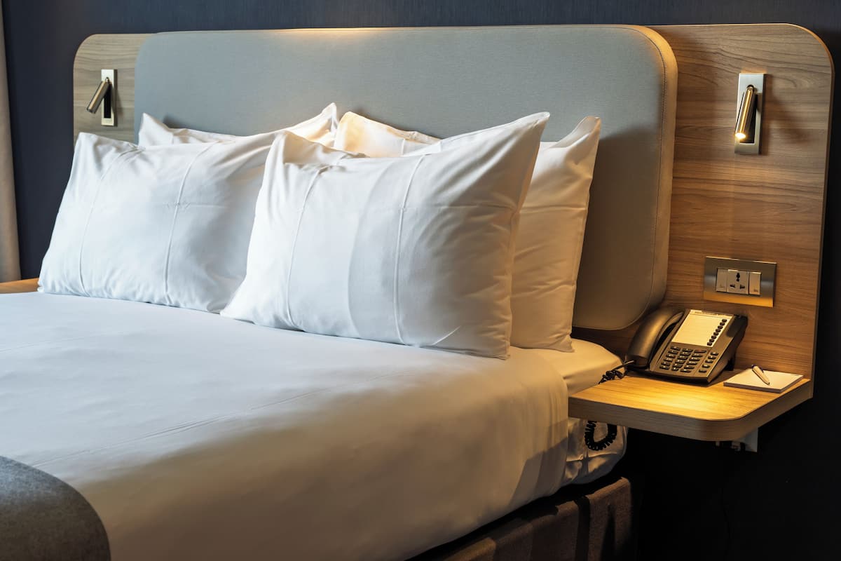 A neatly made bed with white linens and pillows, a bedside table with a telephone and notepad, and a wall-mounted light fixture.