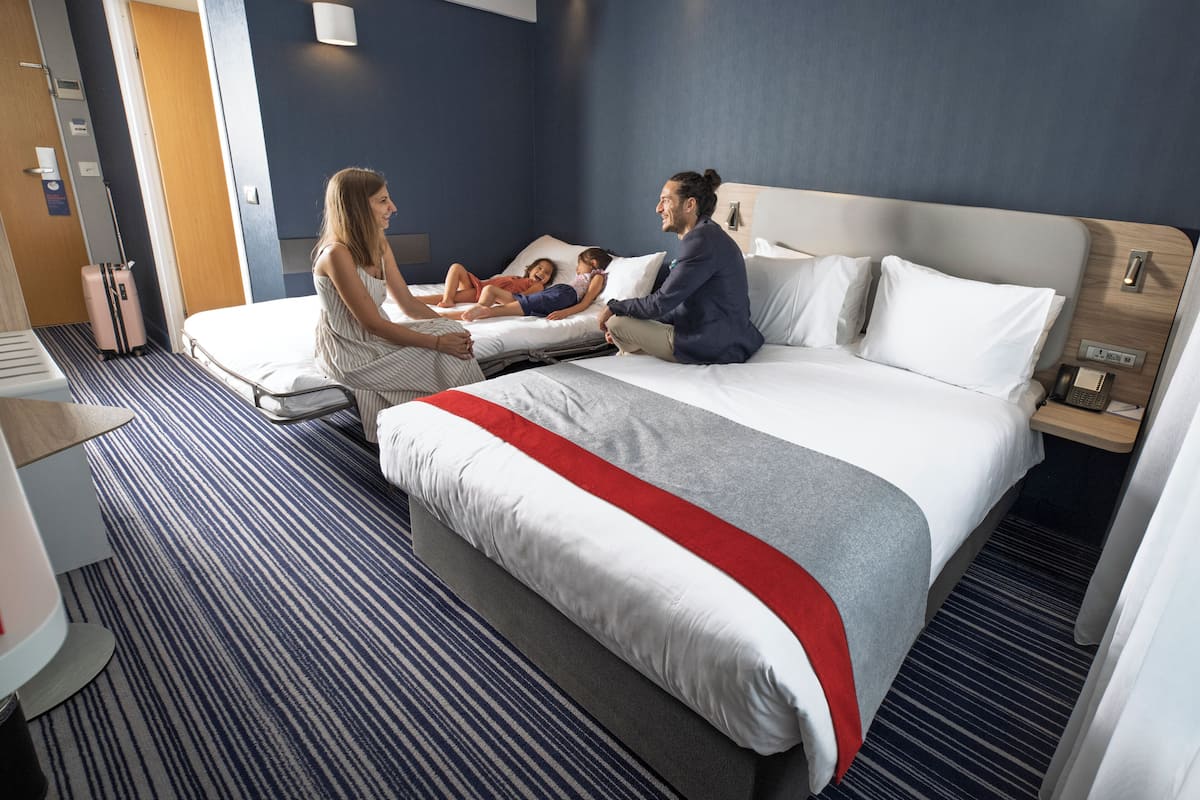 A hotel room with a large bed, a bedside table with a phone, and a suitcase in the corner.