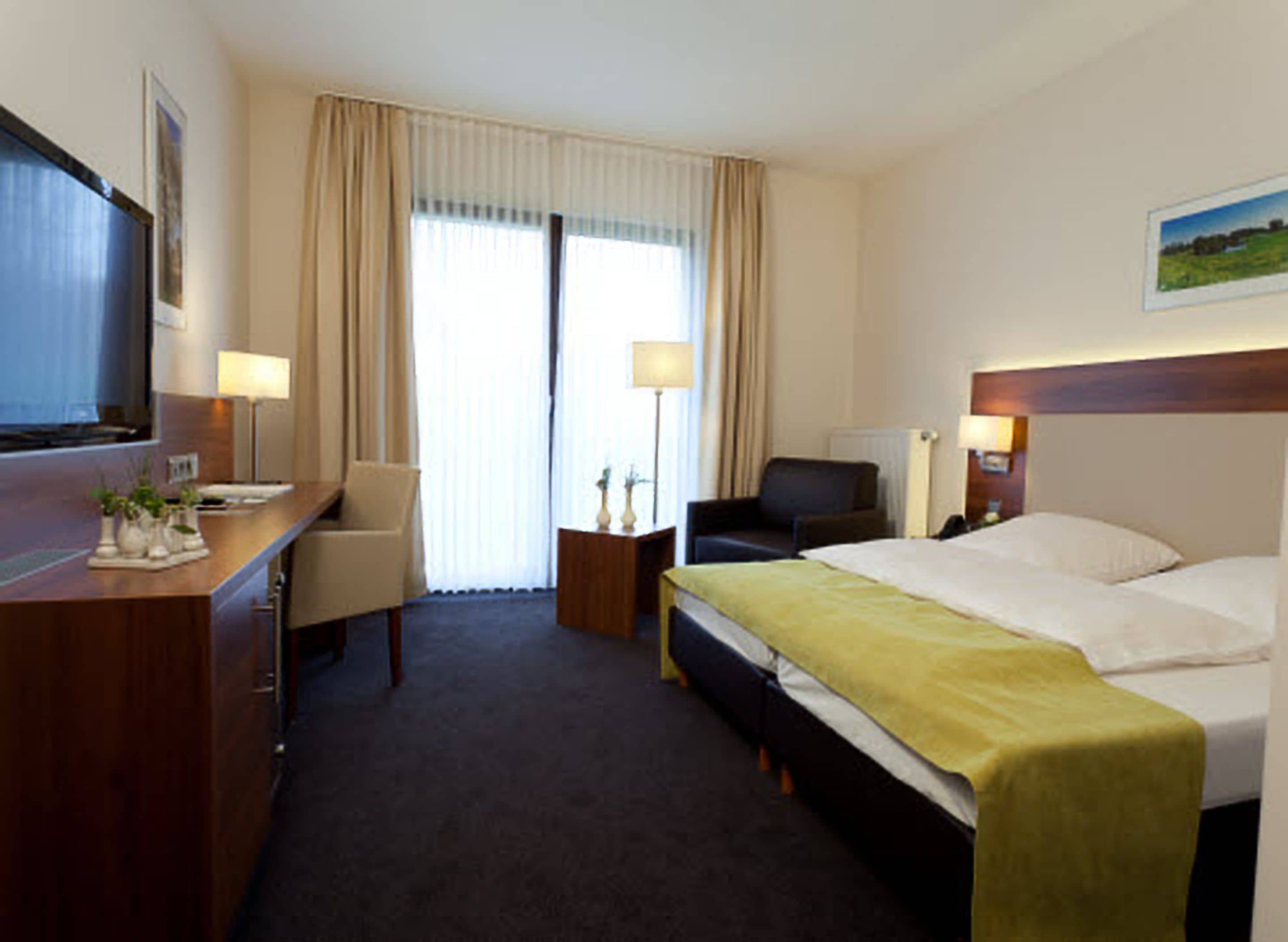 Una habitación de hotel con cama, escritorio, silla, televisor y ventana con cortinas.