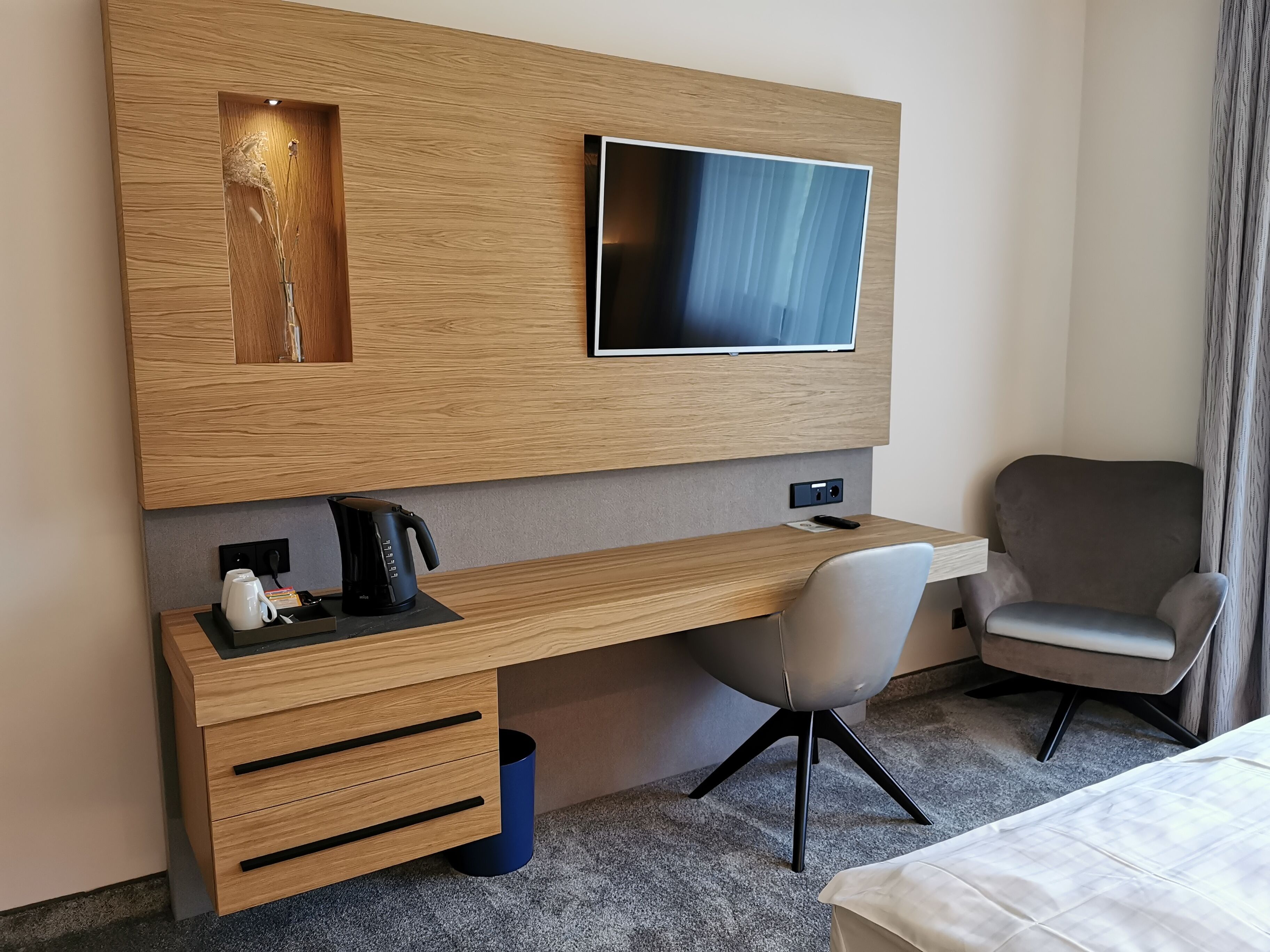 Una habitación de hotel moderna con un escritorio de madera, un televisor, una silla gris y una cama.