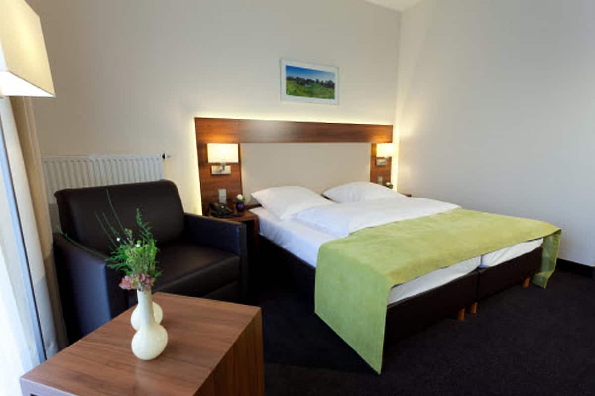 A hotel room with a bed, a chair, a nightstand, a lamp, and a small table with a vase.