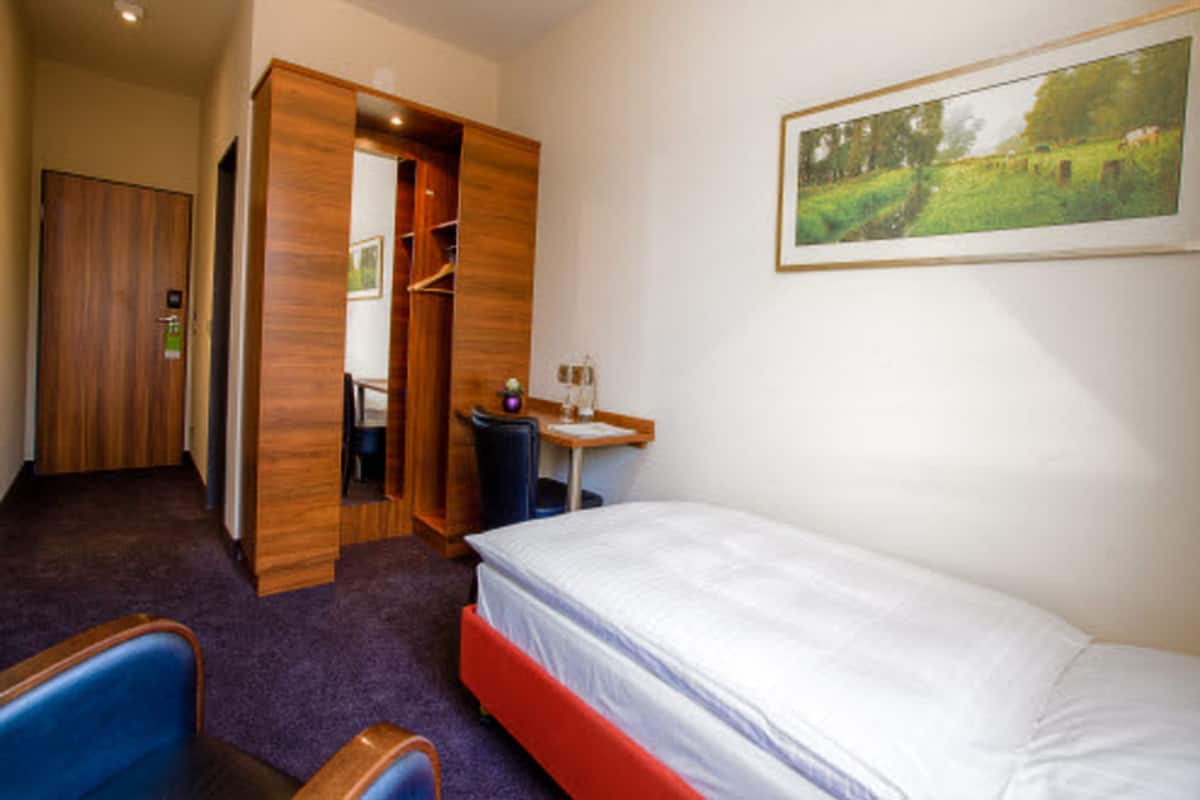 A hotel room with a bed, a desk, a chair, a wardrobe, and a painting on the wall.