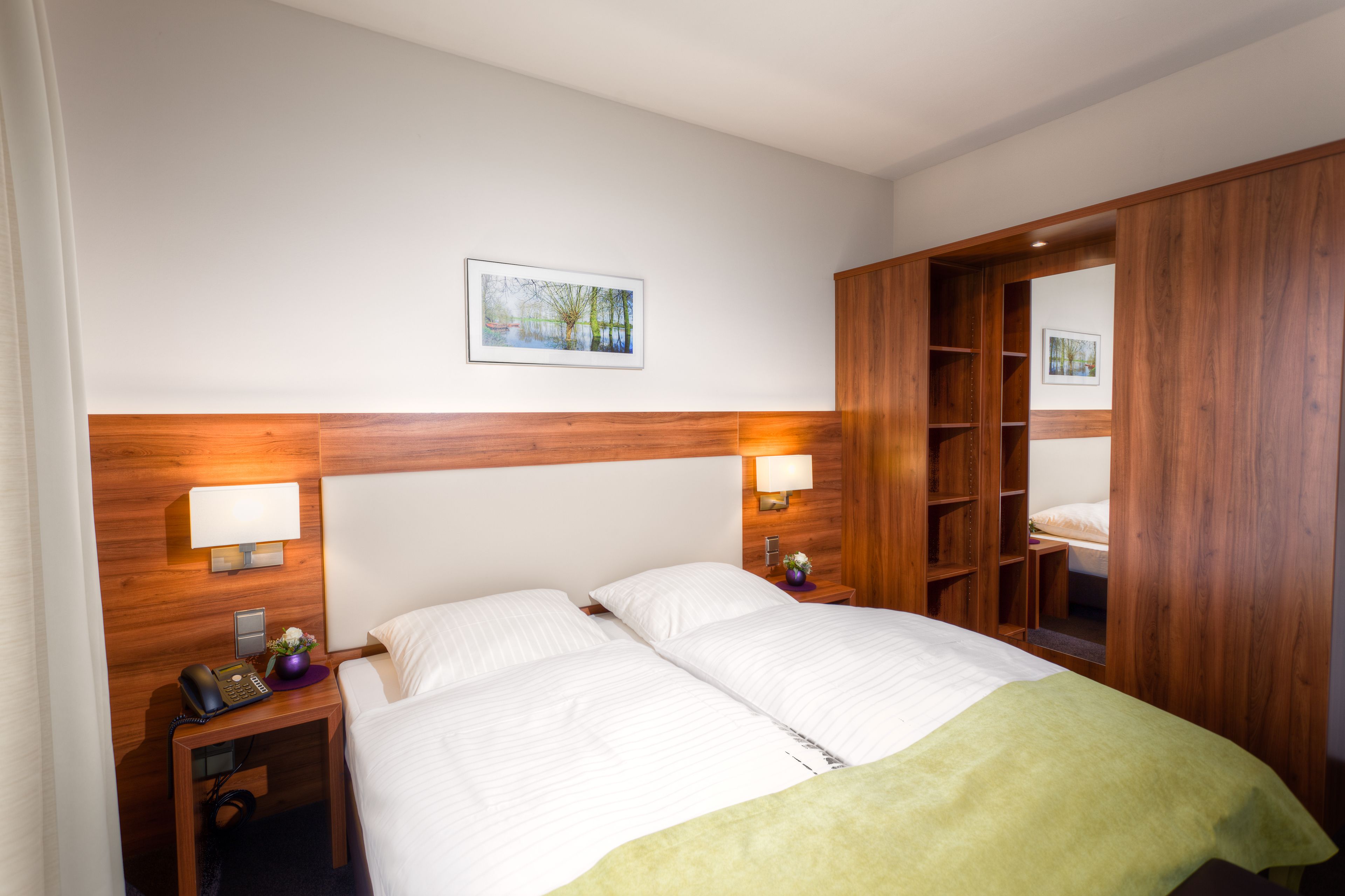 Camera d'albergo con testiera in legno, due letti, lampade sui comodini, un comodino con il telefono, un tavolino con un vaso e un armadio in legno.