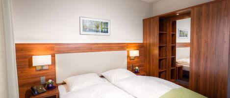 A hotel room with a wooden headboard, two beds, bedside lamps, a nightstand with a phone, a small table with a vase, and a wooden wardrobe.