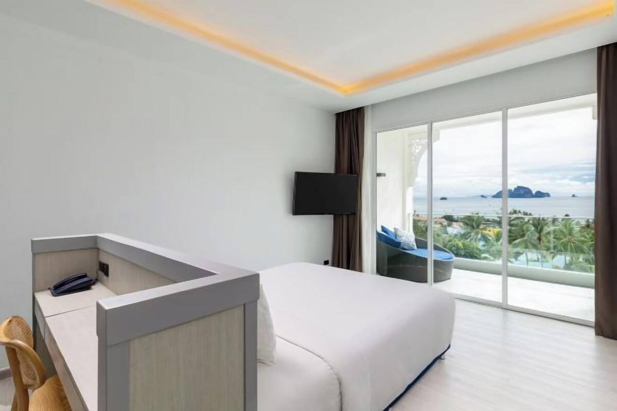 A hotel room with a bed, a desk, a chair, a TV, and a large window with a view of the sea.