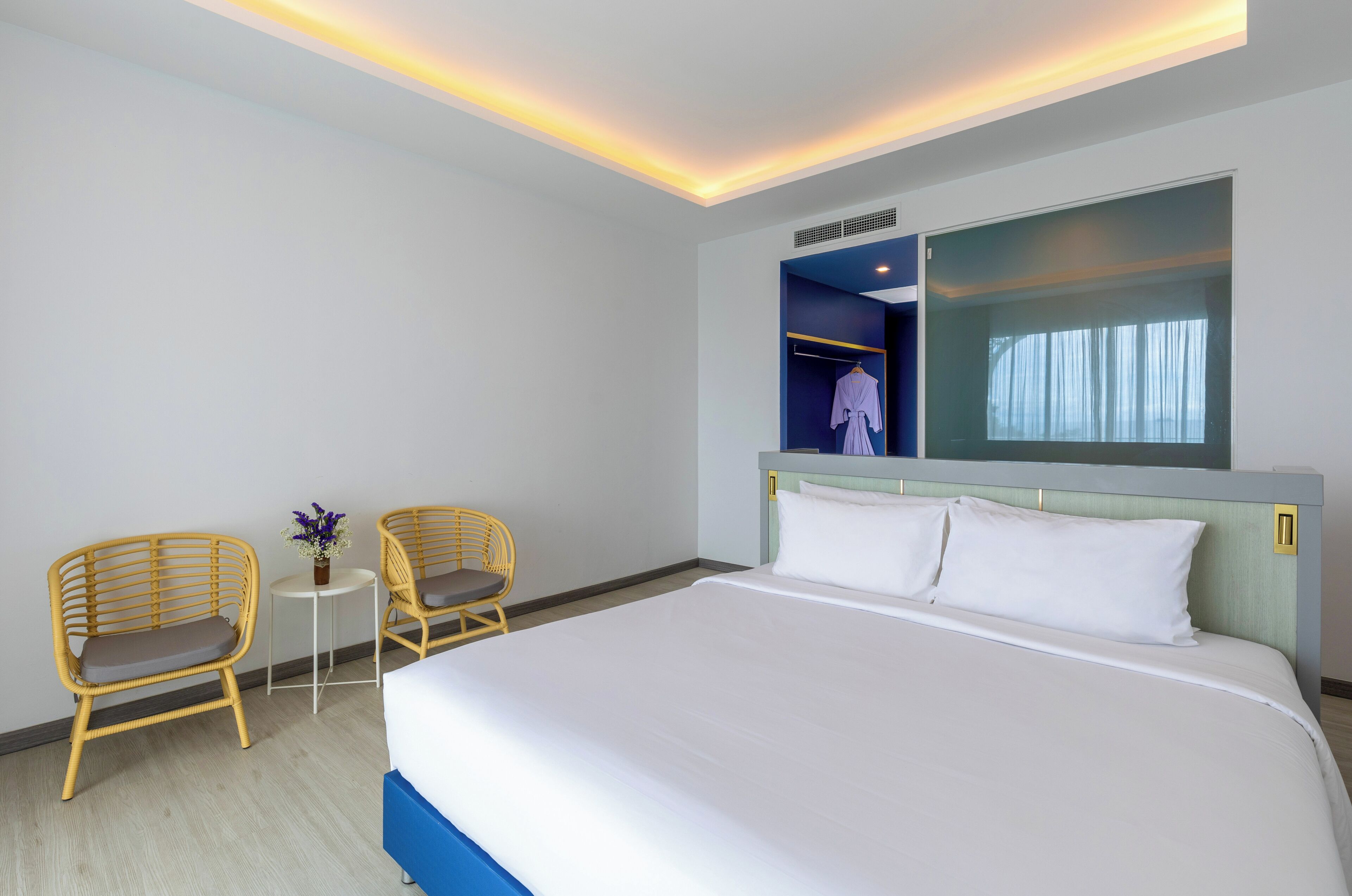 A hotel room with a large bed, two wicker chairs, a small table with a vase, and a mirror above the bed.