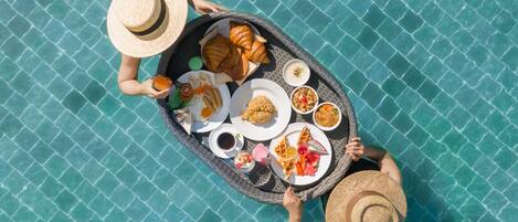 A top-down view of a dining table with various food items and a drink, set on a tiled surface near a pool.