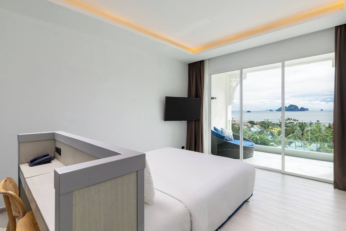Ein modernes Hotelzimmer mit einem großen Bett, einem Schreibtisch mit Telefon, einem Flachbildfernseher und einem Balkon mit Blick auf das Meer und Palmen.
