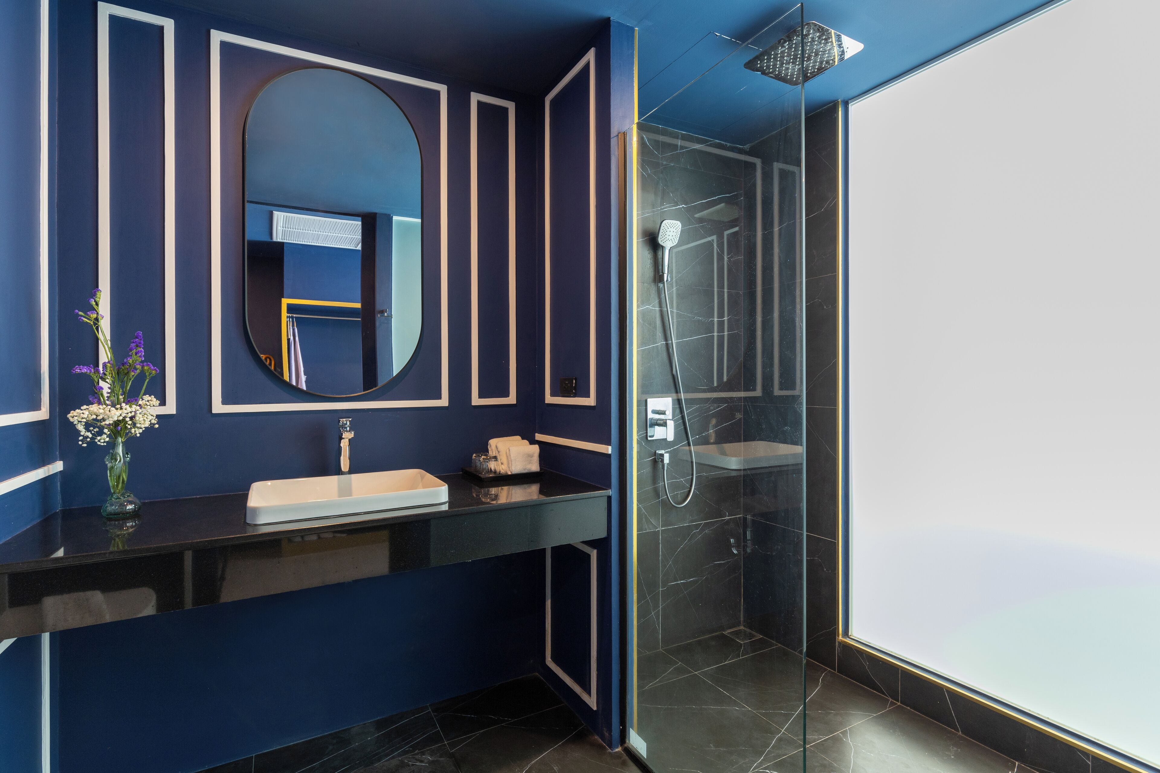 A modern bathroom with a dark blue wall, a round mirror, a black countertop, and a glass-enclosed shower.