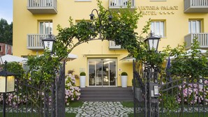 Der Eingang des Viktoria Palace Hotels mit einem steinernen Weg, der zum Eingang führt, umgeben von einem schmiedeeisernen Tor und blühenden Sträuchern.
