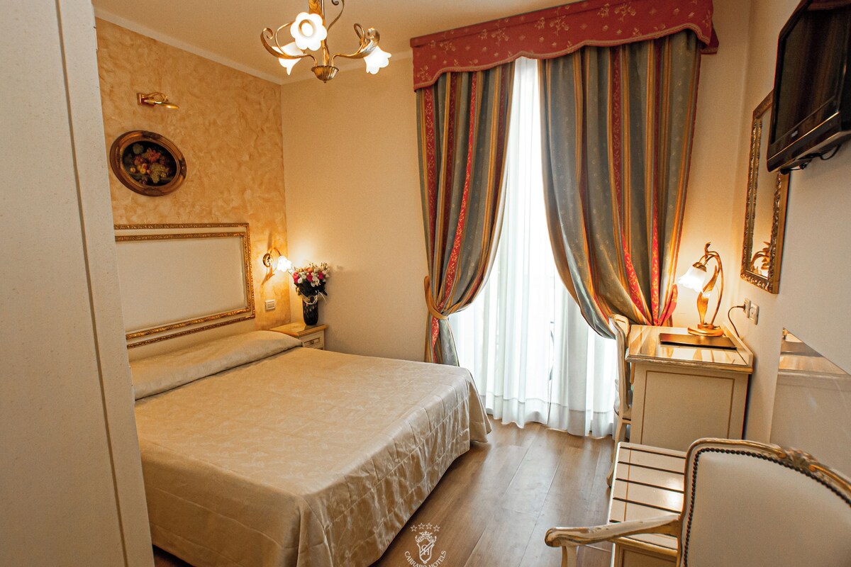 Habitación de hotel con cama, escritorio con lámpara, una silla y televisor en la pared.
