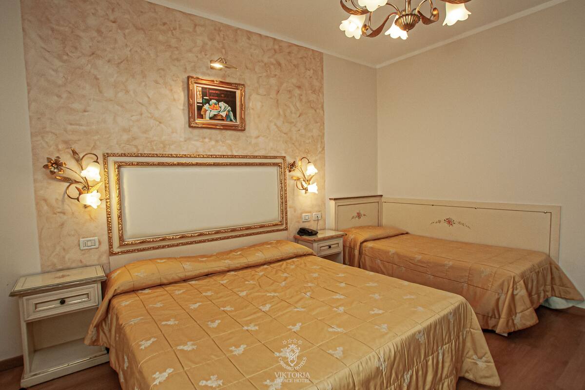 Hotellihuone, jossa on kaksi sänkyä, suuri peili, seinävalaisimet, kattokruunu ja seinällä oleva kehystetty taulu.