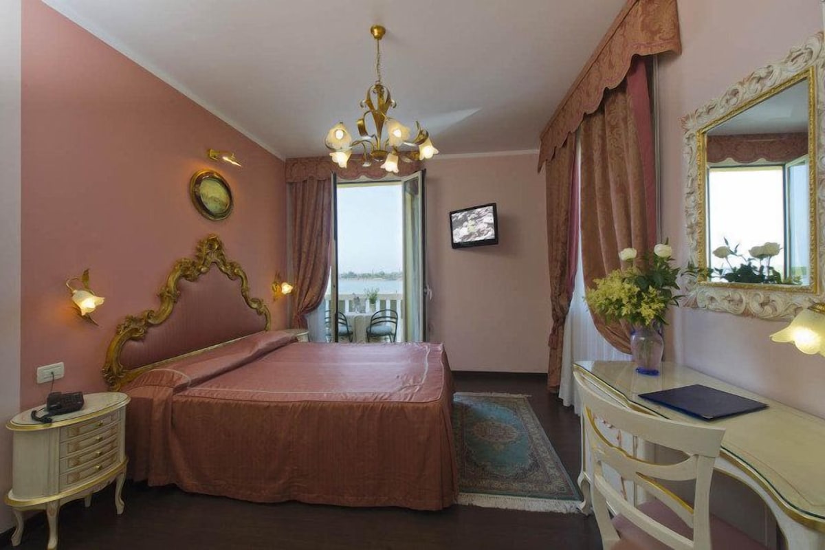 A bedroom with a bed, bedside table, dresser, mirror, chandelier, and a view of the outside.
