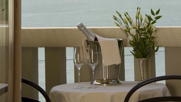 Ein Tisch mit weißer Tischdecke, zwei Weingläsern, einer Serviette und einer Vase mit weißen Blumen, mit Blick auf ein Gewässer und Gebäude im Hintergrund.