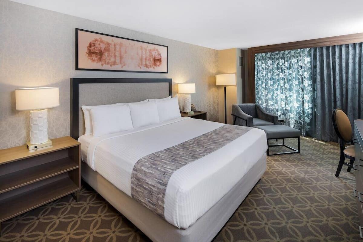 A hotel room with a large bed, two bedside tables with lamps, a chair, a desk, and a window with curtains.