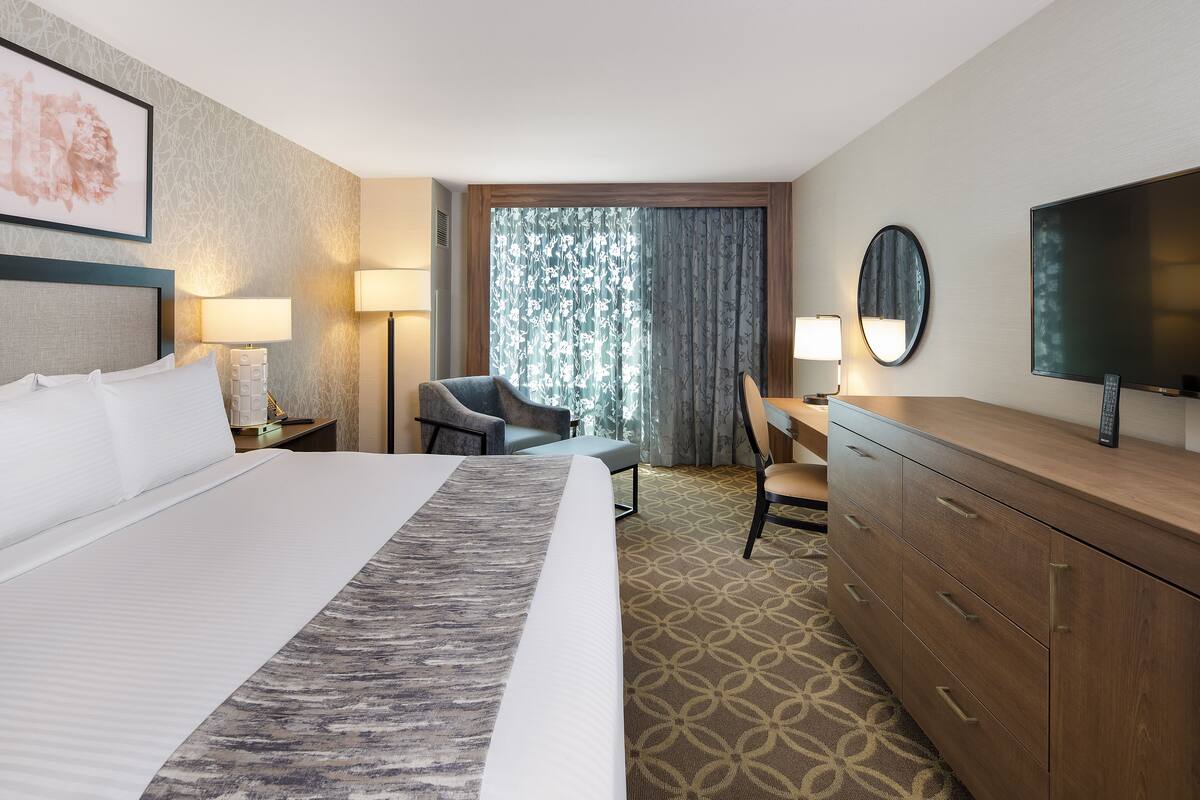 A hotel room with a large bed, a desk, a chair, a TV, and a window with curtains.