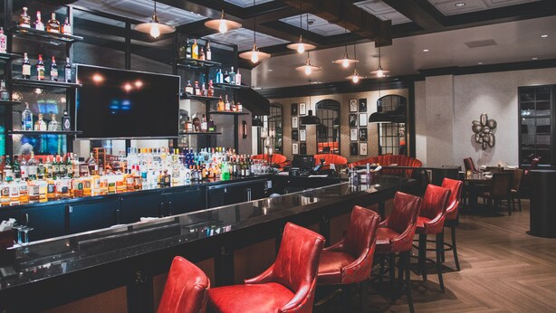 Una zona de bar con una barra larga, múltiples estantes con botellas y taburetes de cuero rojo.