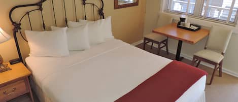 A neatly made bed with white linens and a red throw blanket, a wooden nightstand with a lamp, a small table with a chair, and a window with blinds.