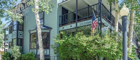 A Victorian-style building with a balcony, American flag, and lush greenery.